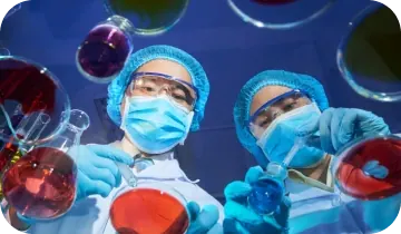 Two lab workers in masks, caps and goggles holding flasks of red and blue liquid